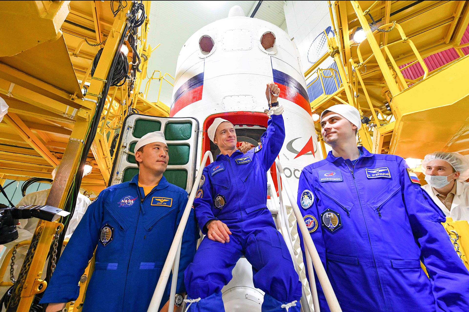 two cosmonauts and an astronaut, all in blue coveralls, pose in front of thier spacecraft while one holds up a small ornament of an angel
